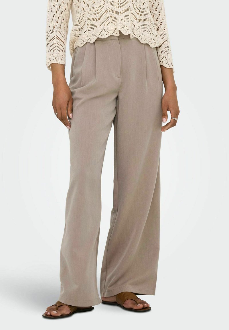 Pantaloni beige a gamba larga indossati con sandali marroni open-toe e un top a maniche lunghe lavorato a uncinetto color crema, mani rilassate ai lati.