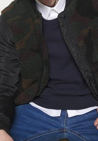 Man som bär blå jeans, vit skjorta med knappar, mörkblå tröja och jacka med kamouflagemönster och svarta ärmar, sittande med händerna vilande.