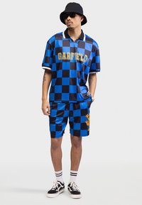 Blå og svart rutete polo- og shortsett med "GARFIELD" i gult. Har et tegneserie-design og hvite detaljer. Bæres med solbriller og joggesko.