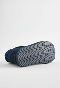 Marinblå mockaskor med en flexibel, texturerad grå gummisula som har ett vågigt mönster. Ingen synlig hårdvara eller detaljer.