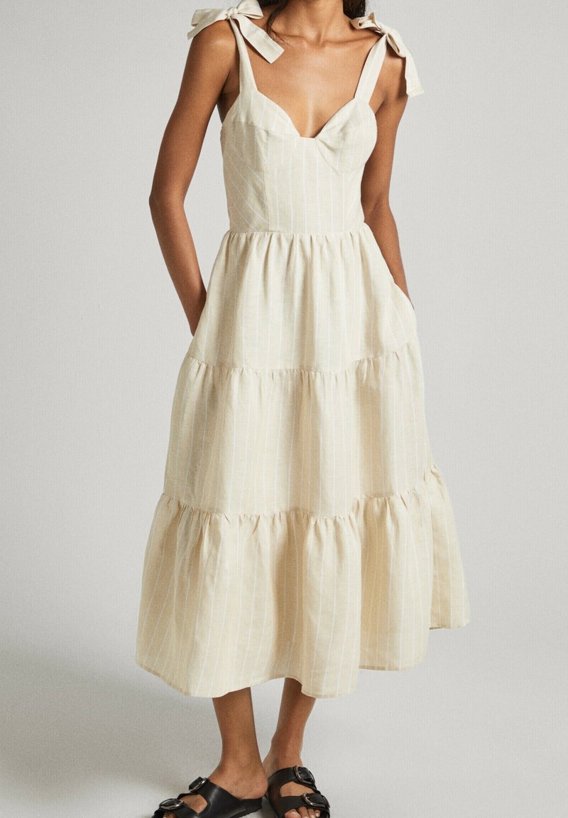Femme portant une robe midi crème sans manches avec liens aux épaules et jupe à volants, associée à des sandales noires à boucle.