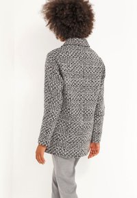 Šedý a černý kostkovaný kabát s vysokým límcem a dlouhými rukávy, vyrobený z texturované tkaniny, s tvarovaným střihem a střední délkou.