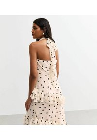 Light beige halter dress with black polka dots, featuring a flowing ruffle hem and a tied bow detail at the back. Smooth, lightweight fabric.