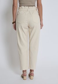 Pantaloni beige a vita alta con cintura arricciata, tasche posteriori e design a gamba dritta. Indossati con sandali a blocco con tacco di colore chiaro.