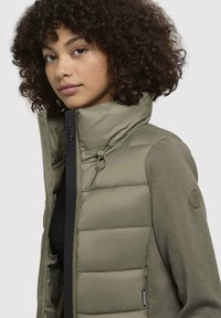 Jachetă izolată verde cu guler înalt, având secțiuni quilted și un material tricotat pe manșete, cu un patch cu logo pe umăr.