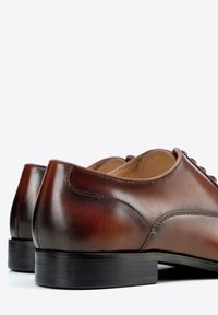 Brązowe skórzane buty wizytowe o eleganckim designie, zakrzywionym obcasie i gładkiej fakturze. Posiadają subtelne przeszycia i płaską czarną podeszwę.