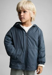 Mornarsko modra jakna s kapuco, sprednjim zadrgo, elastičnimi manšetami in dvema stranskima žepoma. Narejena iz gladkega, lahkega, vodoodpornega materiala.