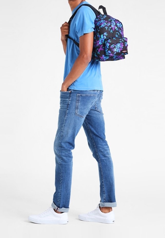 Zwarte rugzak met paarse en blauwe bloemenpatronen, gedragen op één schouder. De persoon draagt een blauwe shirt en opgerolde denim jeans met witte sneakers.