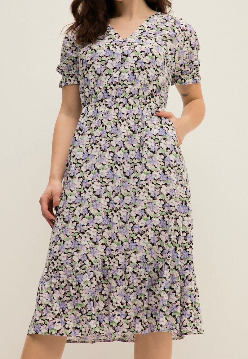 Femme portant une robe fleurie mi-longue avec des manches courtes bouffantes et une taille cintrée, main dans la poche sur un fond uni.