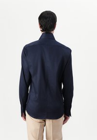 Cămașă bleumarin cu mânecă lungă, cu guler clasic, textură netedă, croială ajustată și tiv cu slit lateral, asortată cu pantaloni bej deschis.