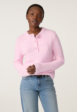 Vrouw met kort haar die een roze pluizige trui en blauwe spijkerbroek draagt, staat met gevouwen handen tegen een effen lichte achtergrond.