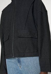 Giacca nera corta con motivo a spina di pesce, chiusura con zip, due tasche frontali e un piccolo etichetta di marca di lato.