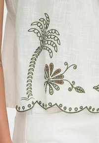 Top de lino blanco con bordado verde de una palmera y motivos florales. El dobladillo con forma de onda y acentos circulares añade detalle.