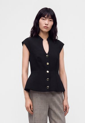 Femme portant un haut noir sans manches boutonné avec un bas peplum et un pantalon gris sur un fond clair uni.