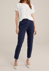 Vit kortärmad topp i kombination med marinblå skräddarsydda byxor och vita spetsiga klackskor. Slätt tyg med subtil lyster, figurnära silhuett.
