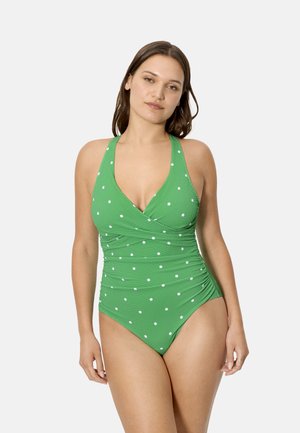 Mujer con traje de baño verde de cuello halter con lunares blancos, de pie frente a un fondo claro y liso.