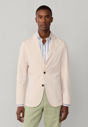 Hombre con blazer beige sobre camisa blanca con rayas azules y pantalones verde claro, de pie frente a un fondo gris.