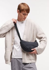 Svart, vadderad påse med en rundad form och ett band, hålls av en person som bär en ljus beige strukturerad kavaj över en vit t-shirt.