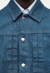 Κοντινή φωτογραφία μπλε τζιν μπουφάν με ασημένια κουμπιά φορεμένο πάνω από ένα λευκό πουκάμισο με γιακά.