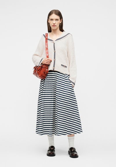 Kvinde stående iført en hvid tekstureret cardigan med marineblå kant, en navy- og hvidstribet midinederdel, hvide sokker, sorte loafers og en rød skuldertaske.