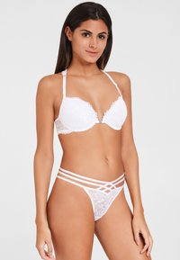 Weißer Spitzen-BH mit Bügel und gewelltem Saum, kombiniert mit einem weißen String, der mit Spitzenverzierungen und einem Kreuzdesign versehen ist.