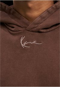 Sudadera con capucha marrón hecha de tela acanalada, con una firma bordada en blanco en la zona del pecho.