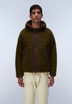 Man draagt een olijfgroene fleecejas met capuchon en beige broek, staand tegen een effen lichte achtergrond.