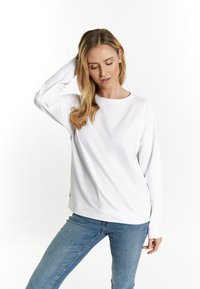 Hanorac alb cu mânecă lungă, de tip crew neckline, cu un croi lejer și mansete cu nervuri, asociat cu blugi albaștri deschis, într-un decor minimalist.
