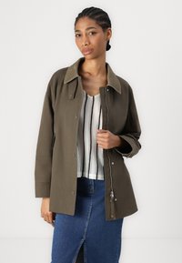 Zelená olivová bunda s měkkým, kontrastním límcem, knoflíky a kapsou na zip. Nositá přes pruhovaný pletený top a džínsovou sukni.