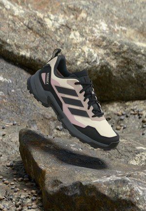 Zapatilla de trail running con detalles en negro, beige y rosa, suspendida sobre terreno rocoso, con suela gruesa y marca "TERREX".