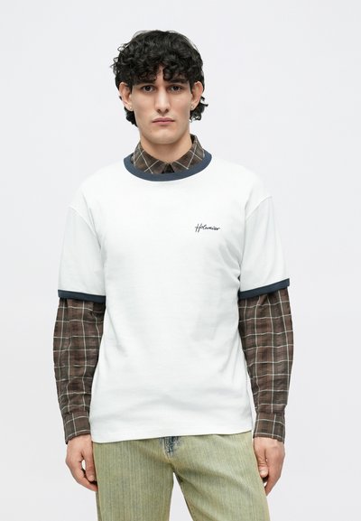 Fehér, rövid ujjú póló tengerészkék bordázott gallérral, barna kockás hosszú ujjú ing fölött rétegezve, világos farmernadrággal párosítva.