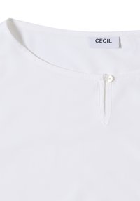 Blouse blanche avec un décolleté rond, dotée d'une fermeture à un bouton et d'une petite fente au col. Tissu doux et lisse.