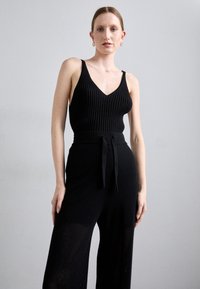 Canotta nera in maglia con scollatura a V e texture a coste, abbinata a pantaloni a gamba larga con vita alta, dotati di una cintura a lacci e un motivo a intreccio aperto.