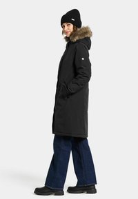 Zwarte geïsoleerde jas met nepbont op de capuchon, gecombineerd met een zwarte brei-beanie en een wijdvallende blauwe jeans. Voorzien van zakken en een knoopsluiting.