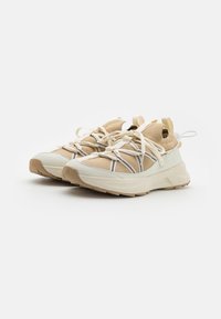 Beżowe sneakersy z teksturowaną cholewą, białymi akcentami i masywną białą podeszwą. Posiadają elastyczne sznurówki i uchwyty do łatwego zakładania.