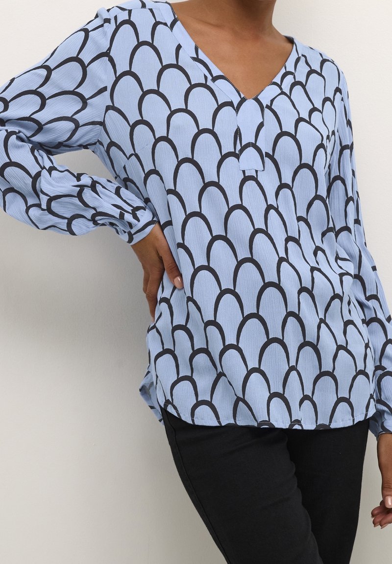 Personne portant un chemisier bleu clair à manches longues avec un motif coquille noir et un pantalon noir, main reposant sur la hanche sur un fond uni.