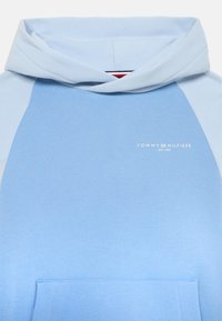 Svetlo modra kapuca z sprednjim žepom, z raglan rokavi in kapuco na vrvico. Na prsih je natisnjen logotip blagovne znamke "TOMMY HILFIGER".