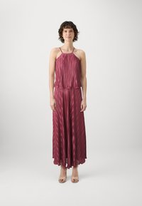 Robe maxi plissée rouge foncé avec des fines bretelles, présentant des rayures verticales et une jupe fluide. Associée à des sandales à talons nudes.