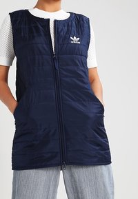 Marinblå vadderad väst med dragkedja fram och två sidofickor. Har Adidas-logotypen på bröstet. Slät textur och kviltad design.