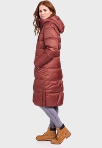 Lange, puffy bordeaux parka met een capuchon, voorzien van een gequiltde textuur, zijzakken en een relaxte pasvorm, gecombineerd met lichtgrijze jeans en bruine laarzen.