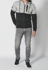 Felpa con zip nera e grigia, con parte superiore texturizzata, cappuccio con lacci, tasca frontale con zip e due tasche laterali, indossata con jeans grigi e sneakers nere.