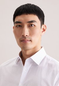 Biała koszula z małymi niebieskimi groszkami, kołnierzyk zapinany na guziki i gładka faktura tkaniny. Modelka ma krótkie czarne włosy i neutralny wyraz twarzy.