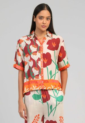 Femme portant une chemise à manches courtes boutonnée et un pantalon assorti avec de grands motifs floraux rouges, blancs et verts sur un tissu crème.