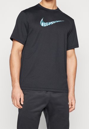 Homme portant un t-shirt noir à manches courtes avec un logo Nike swoosh bleu à motifs et un pantalon noir sur un fond uni.