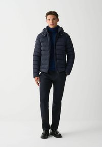 Jūras spēku pufīgs mētelis ar kapuci, ar horizontālu quilted dizainu, ar rāvējslēdzēju un logotipa uzšuvumu kreisajā rokā. Uzvilkts virs augsta kakla krekla.