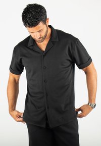 Chemise noire à manches courtes avec un tissu texturé, présentant un col camp, des poignets roulés et six boutons à l'avant.