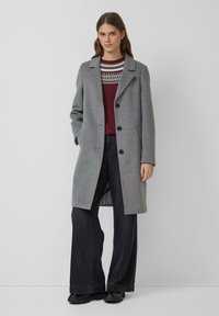 Kobieta ubrana w szary, zapinany na guziki płaszcz, bordowy sweter z białym wzorem, czarne spodnie o szerokich nogawkach i czarne buty, stojąca przy białej ścianie.