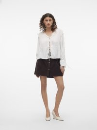 Blouse blanche légère avec des bords volantés et une fermeture à nouer, associée à une jupe en velours côtelé marron avec des boutons sur le devant, et des talons compensés blancs.