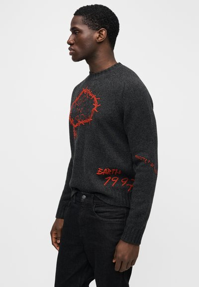 Grå strikket sweater med røde broderede designs, inklusive tekst og et cirkulært mønster, med ribbet halsudskæring og manchetter. Kombineret med sorte jeans.