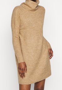 Abito maglione a collo alto beige, realizzato in morbido materiale a maglia. Caratterizzato da maniche lunghe, una vestibilità comoda e un orlo sopra il ginocchio.
